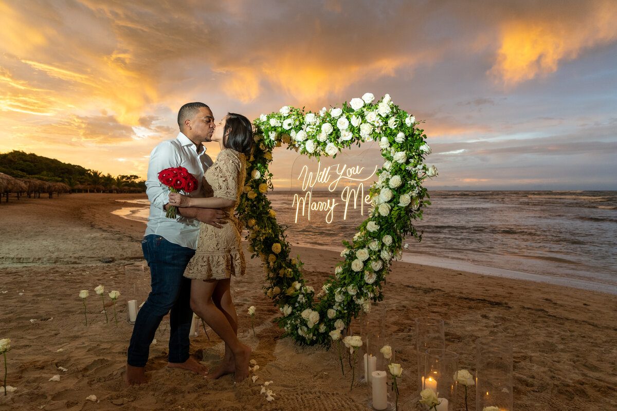 Couple embracing at sunset beside a heart-shaped floral proposal arch